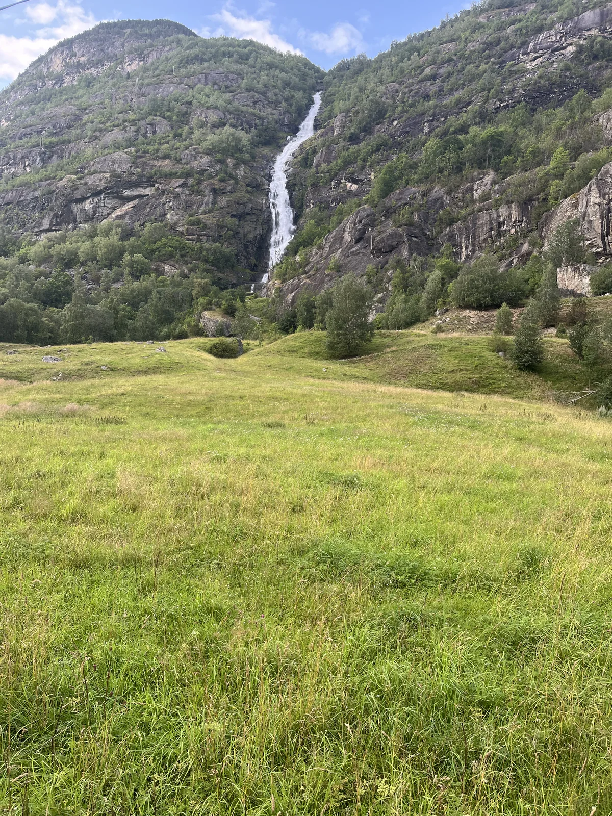 A tall waterfall flowing down the mountainside above a grassy field in Norway